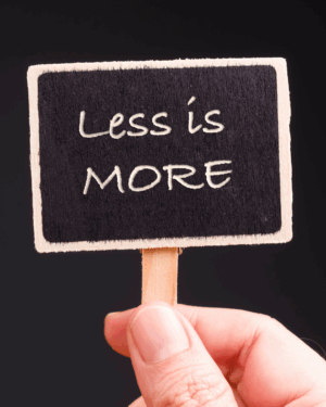 hand holds a small blackboard sign with a wooden border and stick, displaying the phrase "Less is MORE" in white handwritten-style text against a dark background. A hand holds a small blackboard sign with a wooden border and stick, displaying the phrase "Less is MORE" in white handwritten-style text against a dark background. 